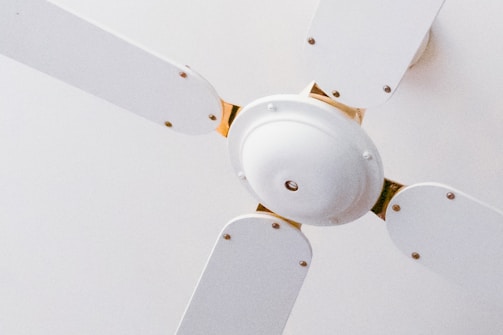 A close-up view of a white ceiling fan with four blades. The fan is mounted on the ceiling with a minimalistic design, featuring metallic screws at the base of each blade. The white color of the fan blends into the ceiling, providing a clean and simple aesthetic.