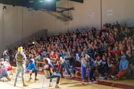A crowded gymnasium with many people seated on bleachers watching an event. Several individuals are actively participating on the gym floor, some of whom are wearing casual clothing and engaging in activities. The atmosphere appears lively and energetic.