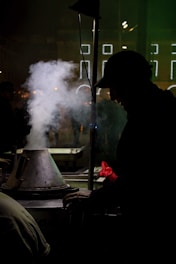 A silhouette of a person, possibly a street vendor, is seen next to a steam-emitting cooking apparatus in a dimly lit urban environment. The background features illuminated rectangular line patterns, suggesting city lights or a decorative installation. The mood is intimate and atmospheric, with hints of warmth from the red glove worn by the vendor.