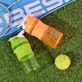 Two colorful plastic bottles, one orange and one green, rest on artificial grass next to a yellow tennis ball and a badminton racket with a blue sports bag partially visible.