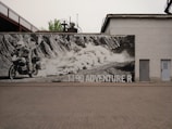 A large black and white mural on a brick wall depicts a motorcyclist riding an adventure bike across a rugged terrain. The rider is kicking up dust as they traverse the landscape. The text '1190 ADVENTURE R' is visible on the mural. The mural is situated outdoors and is surrounded by industrial structures, including a tall building and metal framework.