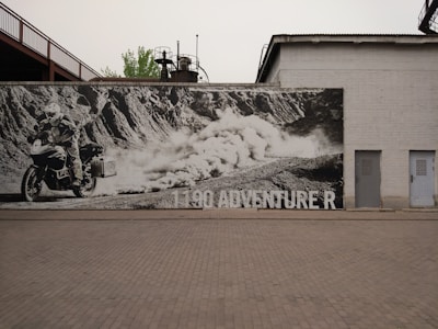 A large black and white mural on a brick wall depicts a motorcyclist riding an adventure bike across a rugged terrain. The rider is kicking up dust as they traverse the landscape. The text '1190 ADVENTURE R' is visible on the mural. The mural is situated outdoors and is surrounded by industrial structures, including a tall building and metal framework.