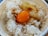 A bowl of white rice topped with a raw egg yolk and drizzled with soy sauce. The rice grains appear fluffy and the egg yolk is vibrant orange, sitting in the center of the bowl. The bowl has a decorative pattern around its edge.