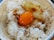 A bowl of white rice topped with a raw egg yolk and drizzled with soy sauce. The rice grains appear fluffy and the egg yolk is vibrant orange, sitting in the center of the bowl. The bowl has a decorative pattern around its edge.