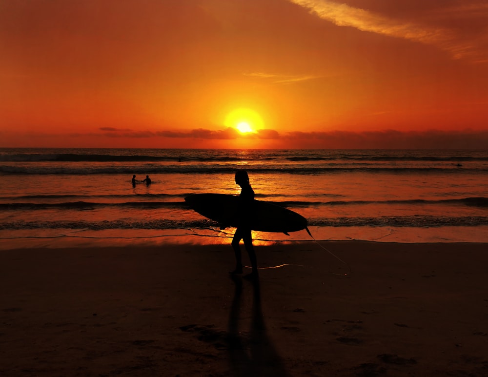 Silhouette eines Mannes, der Surfbrett hält und während des Sonnenuntergangs am Strand spazieren geht