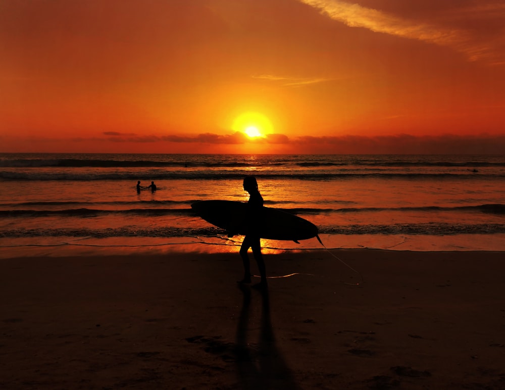 Silueta de hombre sosteniendo tabla de surf caminando en la playa durante la puesta del sol