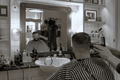 A man is getting a haircut in a barbershop. The scene is reflected in a large mirror that also shows various grooming products on the counter. The barber is standing behind the man, using a clipper and a comb. The ambiance is vintage with tiled walls and a framed photo on a shelf.