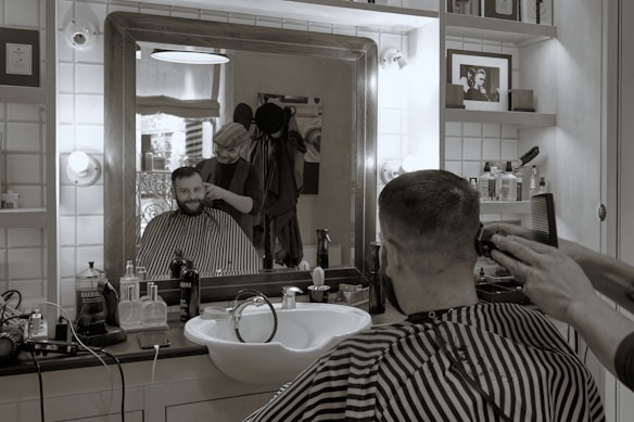 A man is getting a haircut in a barbershop. The scene is reflected in a large mirror that also shows various grooming products on the counter. The barber is standing behind the man, using a clipper and a comb. The ambiance is vintage with tiled walls and a framed photo on a shelf.