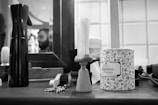 A black and white barbershop scene featuring grooming tools on a counter, including a spray bottle, a shaving brush, a decorative canister labeled 'Talborina Dusting Powder', and a pair of scissors. In the background, a mirror reflects the blurry image of a person with a beard.