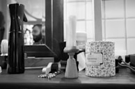 A black and white barbershop scene featuring grooming tools on a counter, including a spray bottle, a shaving brush, a decorative canister labeled 'Talborina Dusting Powder', and a pair of scissors. In the background, a mirror reflects the blurry image of a person with a beard.
