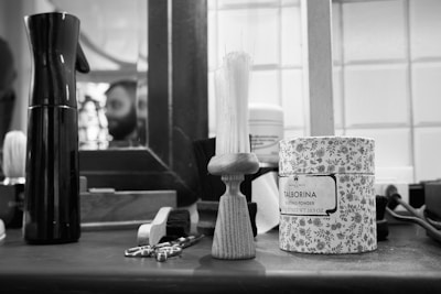 A black and white barbershop scene featuring grooming tools on a counter, including a spray bottle, a shaving brush, a decorative canister labeled 'Talborina Dusting Powder', and a pair of scissors. In the background, a mirror reflects the blurry image of a person with a beard.