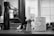 A black and white barbershop scene featuring grooming tools on a counter, including a spray bottle, a shaving brush, a decorative canister labeled 'Talborina Dusting Powder', and a pair of scissors. In the background, a mirror reflects the blurry image of a person with a beard.