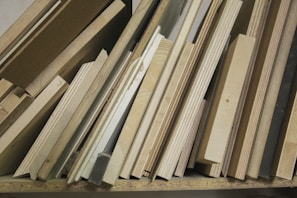 A collection of wooden planks and boards of various sizes, textures, and shades are stacked vertically. The surfaces show grains and patterns typical of wood, ranging from light to medium brown colors. Some boards appear to be plywood, while others might be solid wood pieces.