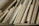 A collection of wooden planks and boards of various sizes, textures, and shades are stacked vertically. The surfaces show grains and patterns typical of wood, ranging from light to medium brown colors. Some boards appear to be plywood, while others might be solid wood pieces.
