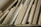 A collection of wooden planks and boards of various sizes, textures, and shades are stacked vertically. The surfaces show grains and patterns typical of wood, ranging from light to medium brown colors. Some boards appear to be plywood, while others might be solid wood pieces.