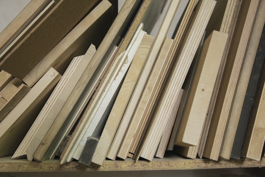 A collection of wooden planks and boards of various sizes, textures, and shades are stacked vertically. The surfaces show grains and patterns typical of wood, ranging from light to medium brown colors. Some boards appear to be plywood, while others might be solid wood pieces.