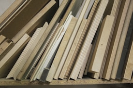 A collection of wooden planks and boards of various sizes, textures, and shades are stacked vertically. The surfaces show grains and patterns typical of wood, ranging from light to medium brown colors. Some boards appear to be plywood, while others might be solid wood pieces.