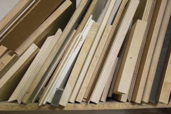 A collection of wooden planks and boards of various sizes, textures, and shades are stacked vertically. The surfaces show grains and patterns typical of wood, ranging from light to medium brown colors. Some boards appear to be plywood, while others might be solid wood pieces.