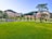 A green lawn stretches across the foreground, bordered by neatly arranged small trees and bushes. In the background, there are academic buildings with modern architecture nestled among taller trees. A row of parked bicycles lines the path, and a few people are casually sitting on the grass, adding a relaxed atmosphere to the scene.