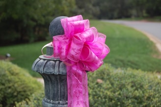 A large, pink ribbon with awareness symbols is tied around a stone or metal post, situated outdoors with greenery and a road in the background.