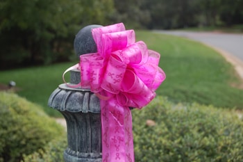 A large, pink ribbon with awareness symbols is tied around a stone or metal post, situated outdoors with greenery and a road in the background.