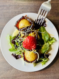 A dish featuring boiled potatoes wrapped in crispy bacon, garnished with fresh green salad leaves and a vibrant red strawberry. Fine sprouts and microgreens are scattered on top. A fork is poised above the food, ready to take a bite.