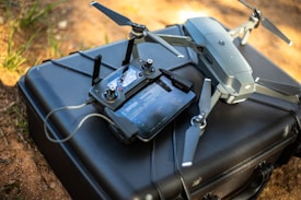 A drone with propellers extended is resting on top of a black carrying case. Next to it is a remote controller connected with a cable, featuring various buttons and a screen displaying an interface. The scene is set outdoors on a surface with some light patches of grass and dirt visible.
