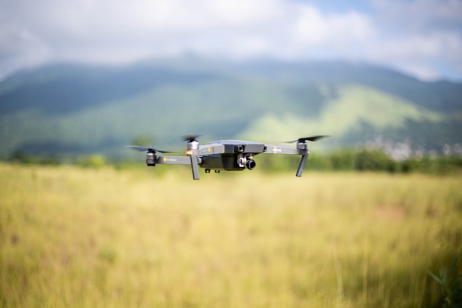A drone is flying over a grassy field with a mountainous landscape in the background. The sky is partly cloudy, adding depth to the serene outdoor scene.