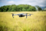 A drone hovers in the air amidst a grassy field with a backdrop of green trees and a slightly cloudy sky. The focus is on the drone, with its camera and propellers clearly visible.