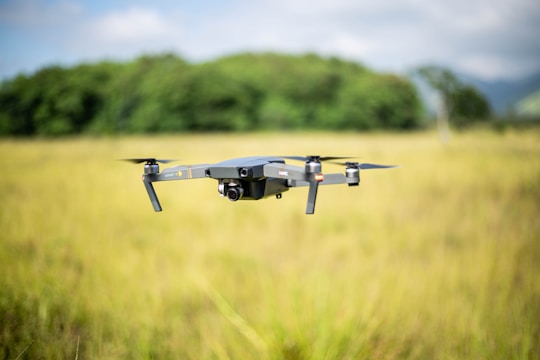 A drone hovers in the air amidst a grassy field with a backdrop of green trees and a slightly cloudy sky. The focus is on the drone, with its camera and propellers clearly visible.