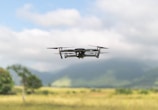 A drone is hovering in the air over a picturesque landscape with a grassy field and distant, fog-shrouded mountains. The drone appears to be modern and sleek, with four propellers visible. The sky is partially cloudy, casting soft shadows over the scene.