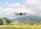 A drone is hovering in the air over a picturesque landscape with a grassy field and distant, fog-shrouded mountains. The drone appears to be modern and sleek, with four propellers visible. The sky is partially cloudy, casting soft shadows over the scene.