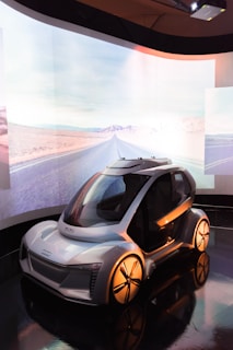 A futuristic concept car displayed in a sleek indoor environment with a large, curved screen in the background showing a desert road. The car features a compact design with smooth curves, large wheels, and a transparent dome-like roof.
