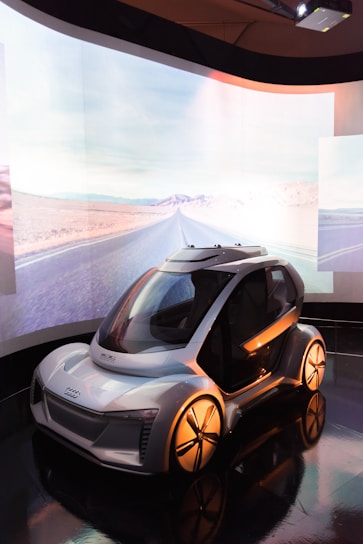 A futuristic concept car displayed in a sleek indoor environment with a large, curved screen in the background showing a desert road. The car features a compact design with smooth curves, large wheels, and a transparent dome-like roof.