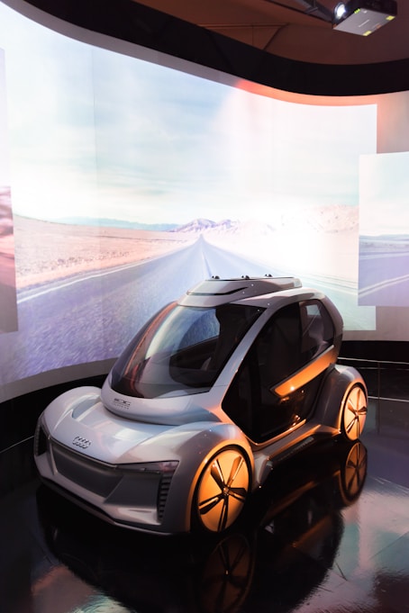 A futuristic concept car displayed in a sleek indoor environment with a large, curved screen in the background showing a desert road. The car features a compact design with smooth curves, large wheels, and a transparent dome-like roof.