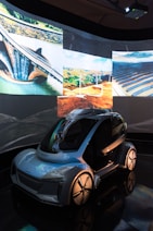 A futuristic, sleek vehicle with a transparent roof and aerodynamic design is displayed in a dimly lit environment. Surrounding the vehicle are projections of eco-friendly technology, including wind turbines, solar panels, and a dam. These images suggest themes of sustainability and innovation.