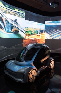 A futuristic, sleek vehicle with a transparent roof and aerodynamic design is displayed in a dimly lit environment. Surrounding the vehicle are projections of eco-friendly technology, including wind turbines, solar panels, and a dam. These images suggest themes of sustainability and innovation.