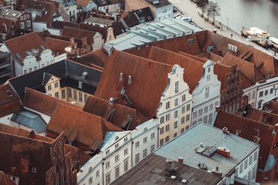 Aerial view of an urban area featuring numerous historic rooftops with red clay tiles and elaborate architectural details like gables and dormers. The scene includes a variety of old buildings tightly packed together, with some modern structures in between. A waterway is visible in the upper part of the image with some trees lining its edge.