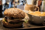 A close-up view of a gourmet burger with a whole grain bun, topped with lettuce and cheese, placed on a rectangular plate. To the right, there is a white bowl filled with golden French fries. The setting appears to be a restaurant with a warm, cozy atmosphere.