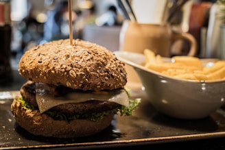 A close-up view of a gourmet burger with a whole grain bun, topped with lettuce and cheese, placed on a rectangular plate. To the right, there is a white bowl filled with golden French fries. The setting appears to be a restaurant with a warm, cozy atmosphere.