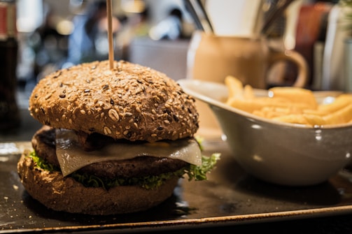 A close-up view of a gourmet burger with a whole grain bun, topped with lettuce and cheese, placed on a rectangular plate. To the right, there is a white bowl filled with golden French fries. The setting appears to be a restaurant with a warm, cozy atmosphere.