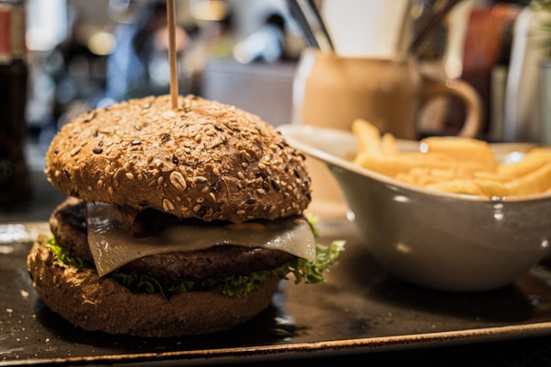 A close-up view of a gourmet burger with a whole grain bun, topped with lettuce and cheese, placed on a rectangular plate. To the right, there is a white bowl filled with golden French fries. The setting appears to be a restaurant with a warm, cozy atmosphere.