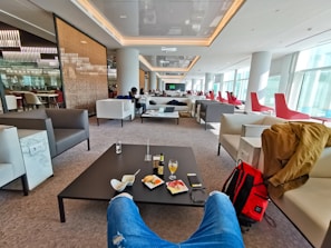 A modern lounge area with comfortable seating, including white chairs and red high-back chairs. People are seated throughout, some engaging with screens or looking out large windows that let in ample natural light. In the foreground, there are food items and a drink on a table, and a red backpack beside a coat on a chair.