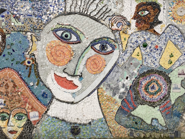 A vibrant mosaic artwork featuring abstract human figures with exaggerated facial features, including large spiral eyes and colorful, textured patterns. The piece combines various small tiles in blues, whites, oranges, and greens to create a dynamic and whimsical aesthetic.