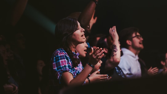 A group of people is gathered in a lively environment, with individuals clapping and appearing to be engaged or cheering. The scene suggests a concert or live event atmosphere.
