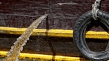A rubber hose is pouring water over the side of a structure with a dark red, textured surface. A black tire, used as a bumper or fender, is suspended to the right with a rope. A thick yellow line runs horizontally across the structure, providing contrast against the darker background.