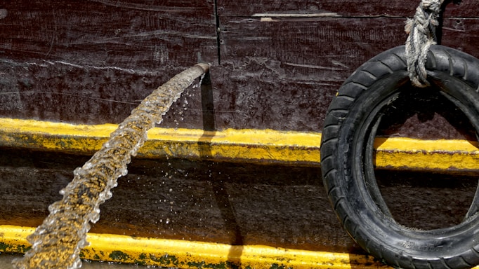 A rubber hose is pouring water over the side of a structure with a dark red, textured surface. A black tire, used as a bumper or fender, is suspended to the right with a rope. A thick yellow line runs horizontally across the structure, providing contrast against the darker background.