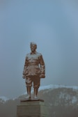 A statue of a uniformed figure stands prominently on a pedestal. The background features misty mountains partially covered with snow, creating a serene and somber atmosphere.