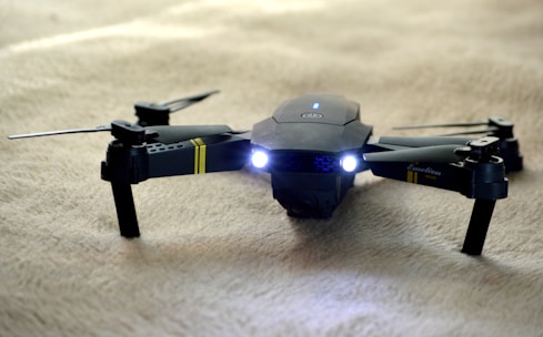 A sleek black drone with extended propeller arms and illuminated front lights is positioned on a soft beige surface. The drone has markings in yellow and is detailed with inscriptions on one arm.