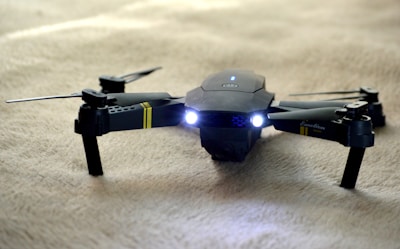 A sleek black drone with extended propeller arms and illuminated front lights is positioned on a soft beige surface. The drone has markings in yellow and is detailed with inscriptions on one arm.